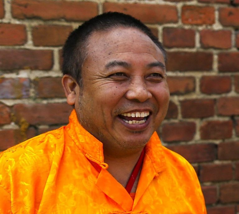 Portrait of Rinpoche laughing, wearing traditional saffron-orange robes, in front of a red brick wall